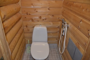 A wooden cabin-style bathroom with a toilet, a shower, and a toilet brush. - Cottage H62 Ruokolahti (Ruokolahti)