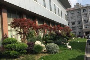 Un edifici amb façana moderna, un jardí ben cuidat amb arbustos diversos i una estàtua blanca, i un edifici clàssic al fons.