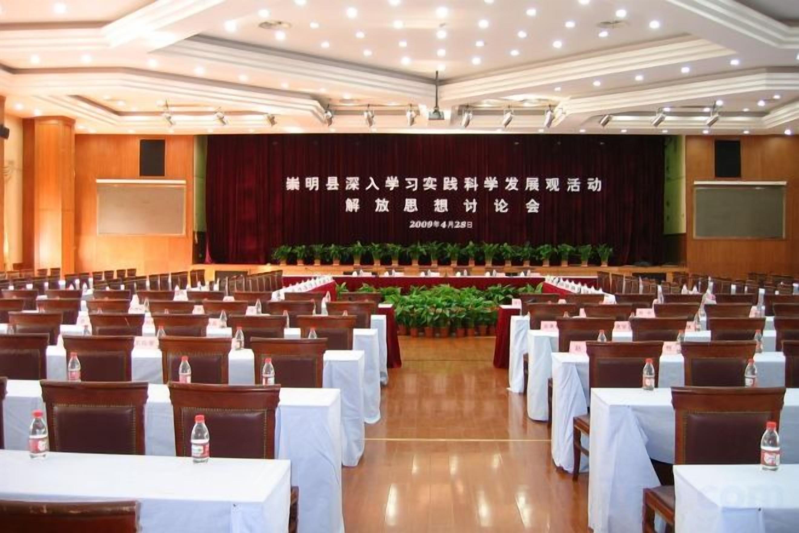 Un salón de conferencias con filas de mesas y sillas, un escenario con una pantalla de presentación y un cartel con texto.