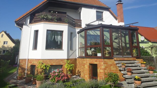 Une maison à deux étages, avec un toit recouvert de tuiles rouges, une véranda en verre et une façade en briques.