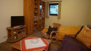 Un salon chaleureux avec un téléviseur, un meuble de rangement en bois, un fauteuil en osier, un canapé et une fenêtre avec des rideaux.