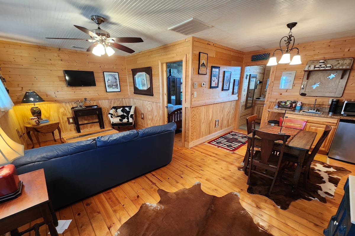 A cozy cabin interior with a wooden floor, a blue sofa, a cowhide rug, a dining area, and a kitchen with a refrigerator.