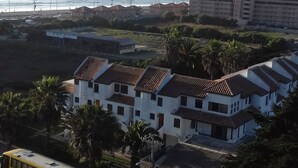 Un hotel frente al mar con varias edificaciones blancas, una carretera visible con un autobús verde y una vista despejada del océano.