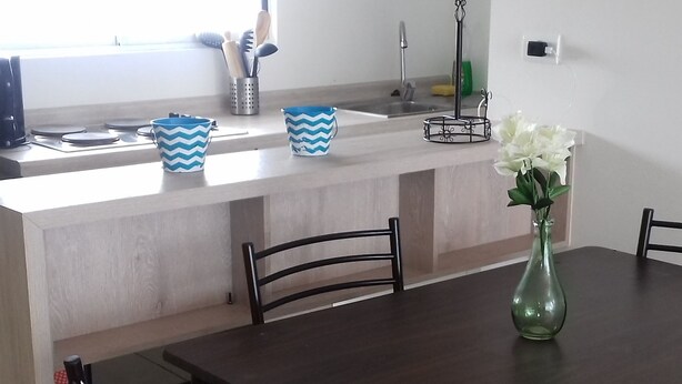Un comedor con una mesa y sillas de madera, una cocina al fondo y un florero con flores sobre la mesa.
