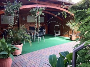 An outdoor patio area with a brick floor, potted plants, a table with chairs, and a wooden structure in the background. - Pension Borna (Borna)