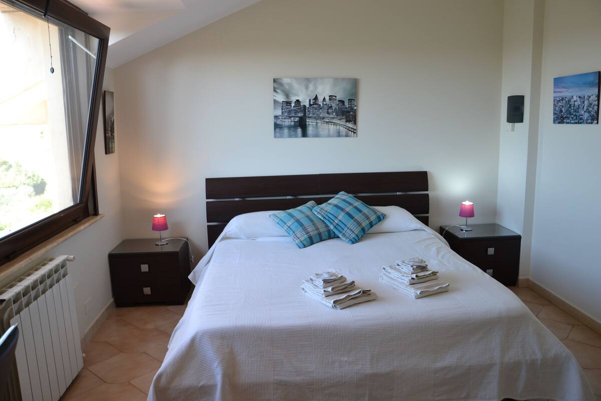 A neatly made bed with a cityscape painting above it, two bedside tables with lamps, and a window with curtains.