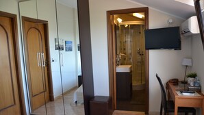 Habitación de hotel con cama, escritorio, silla, televisión, espejo y baño visible a través de una puerta abierta.