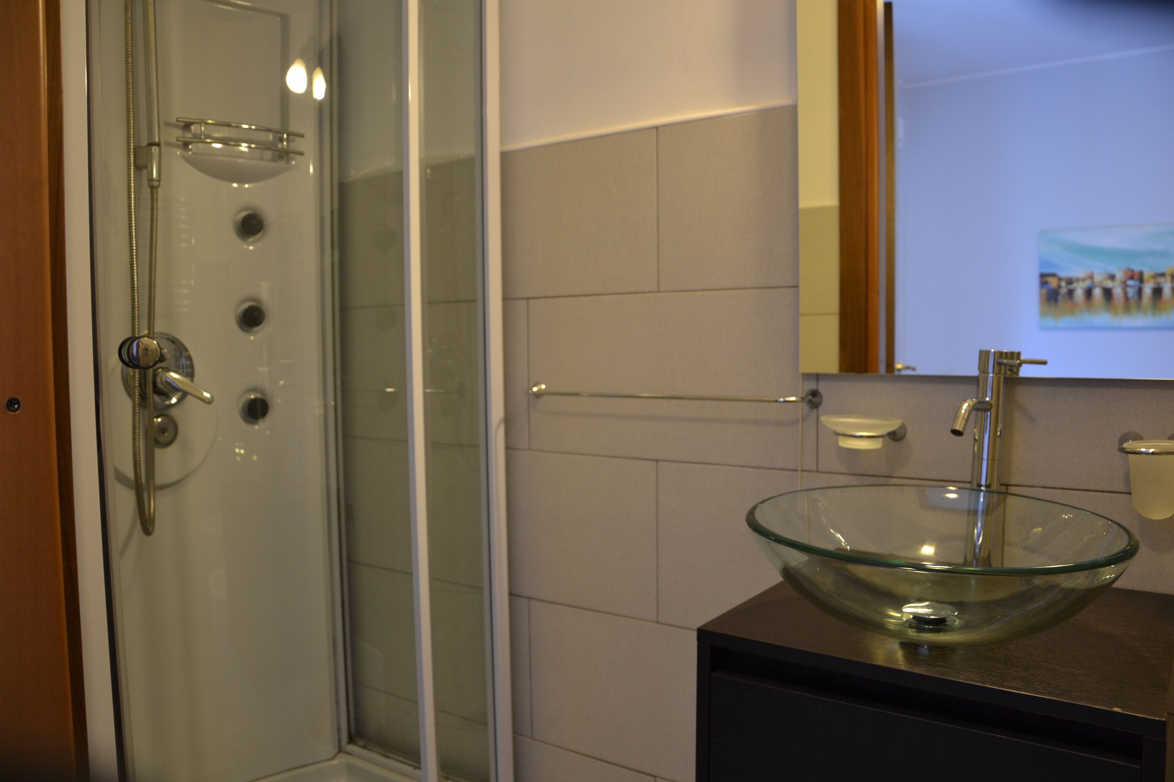 Un baño con lavamanos de cuenco de vidrio, ducha con puerta de vidrio y espejo con un cuadro.