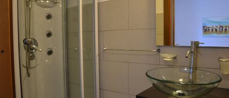 Un baño con lavamanos de cuenco de vidrio, ducha con puerta de vidrio y espejo con un cuadro.