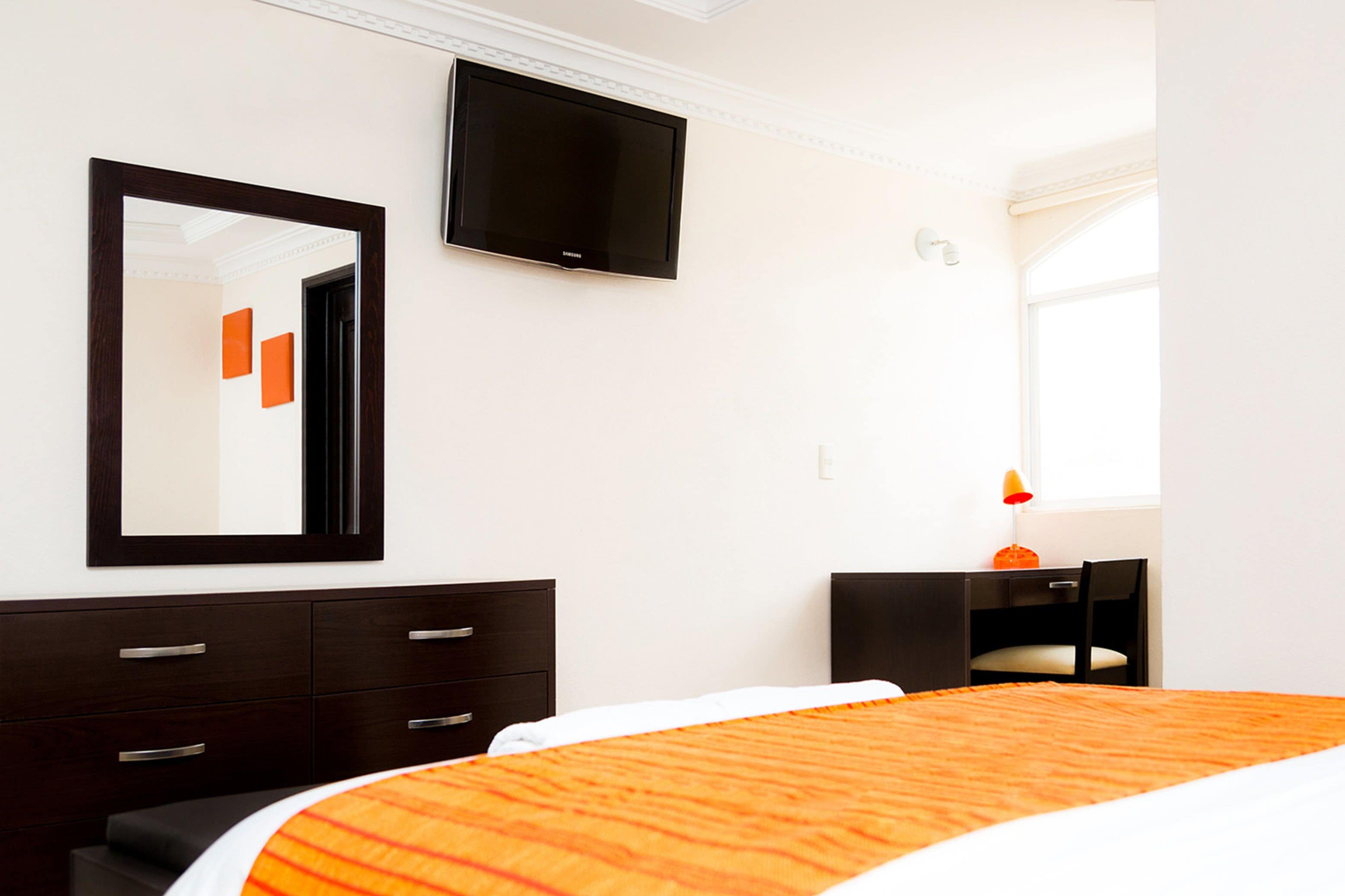 A hotel room with a bed, a TV mounted on the wall, a desk with a chair, and a window.