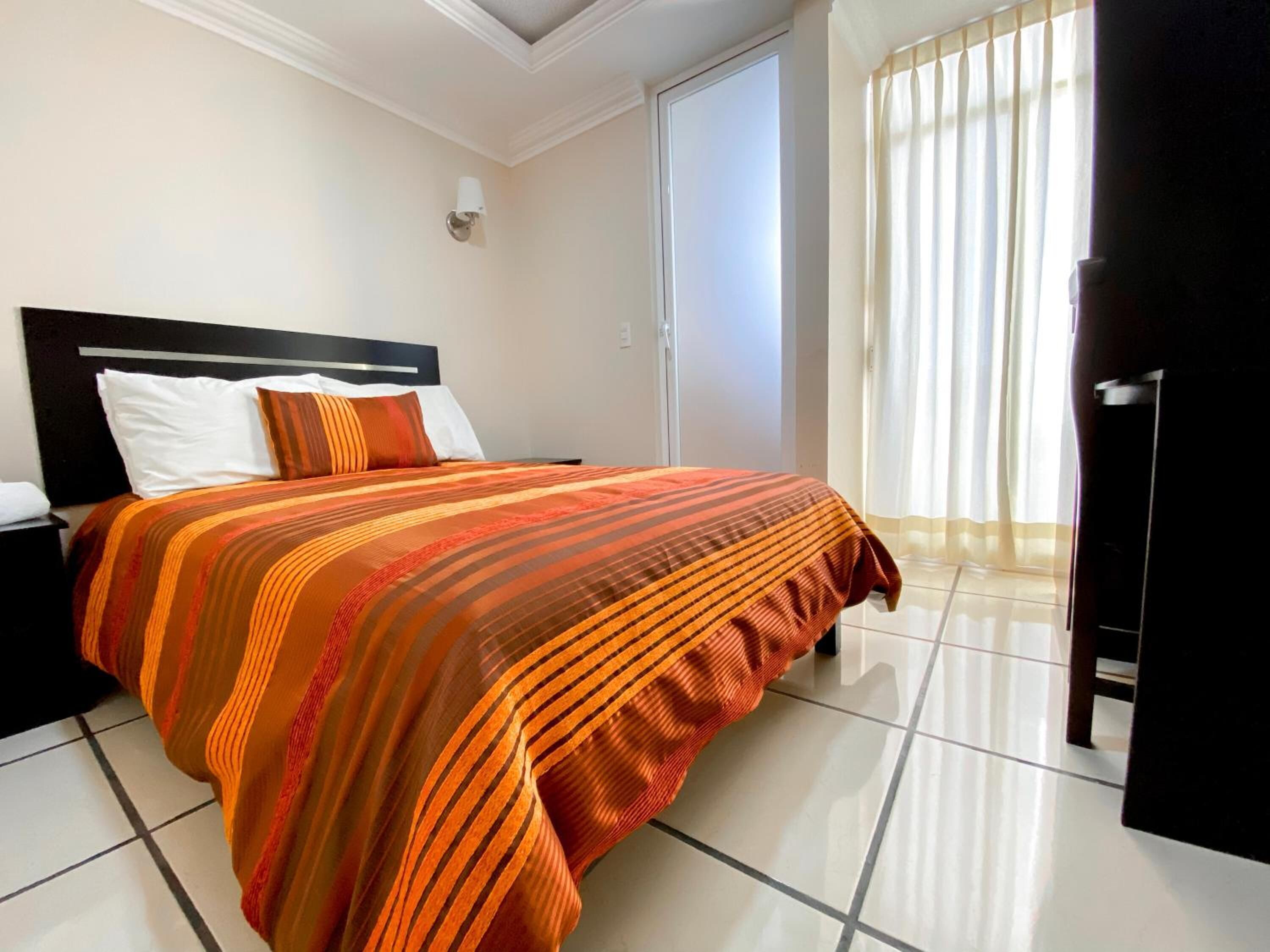 A hotel room with a bed, a nightstand, a wall-mounted lamp, a door, and a window with curtains.