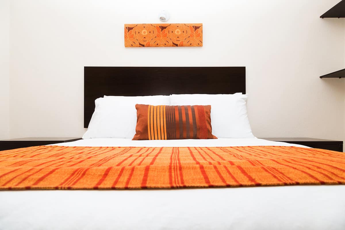 A hotel room with a bed, white bedding, an orange striped blanket, and a wall-mounted artwork.