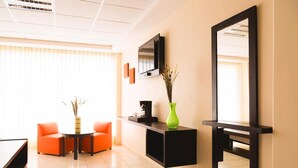 Une chambre d’hôtel moderne équipée d’un canapé, d’une table basse, d’un téléviseur fixé au mur et d’un vase vert contenant des plantes séchées.