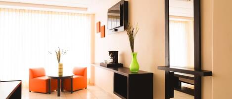 A modern hotel room with a sofa, a coffee table, a TV mounted on the wall, and a green vase with dried plants.