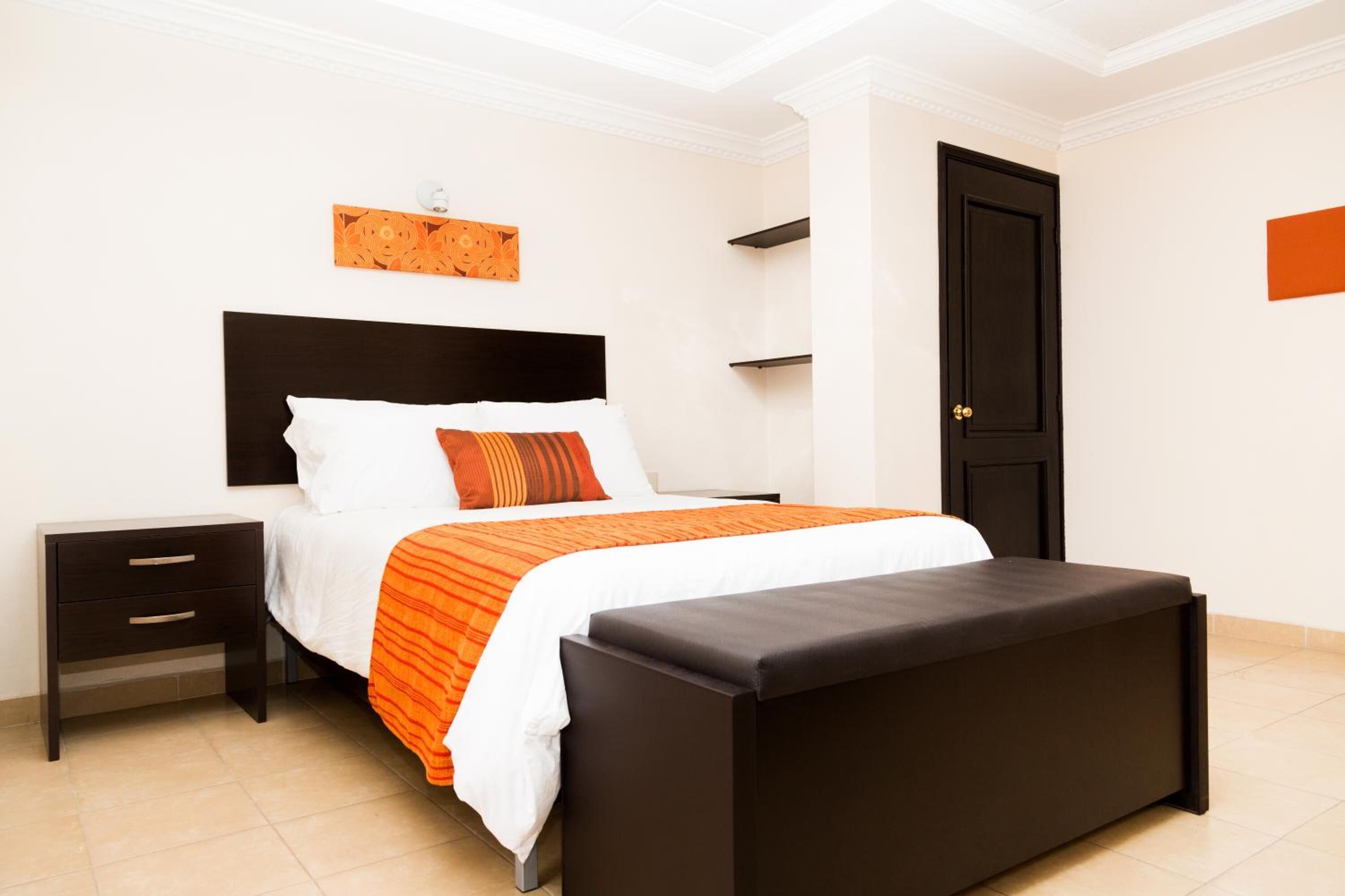 A bedroom with a bed, a nightstand, a bench, a door, and a wall-mounted shelf.