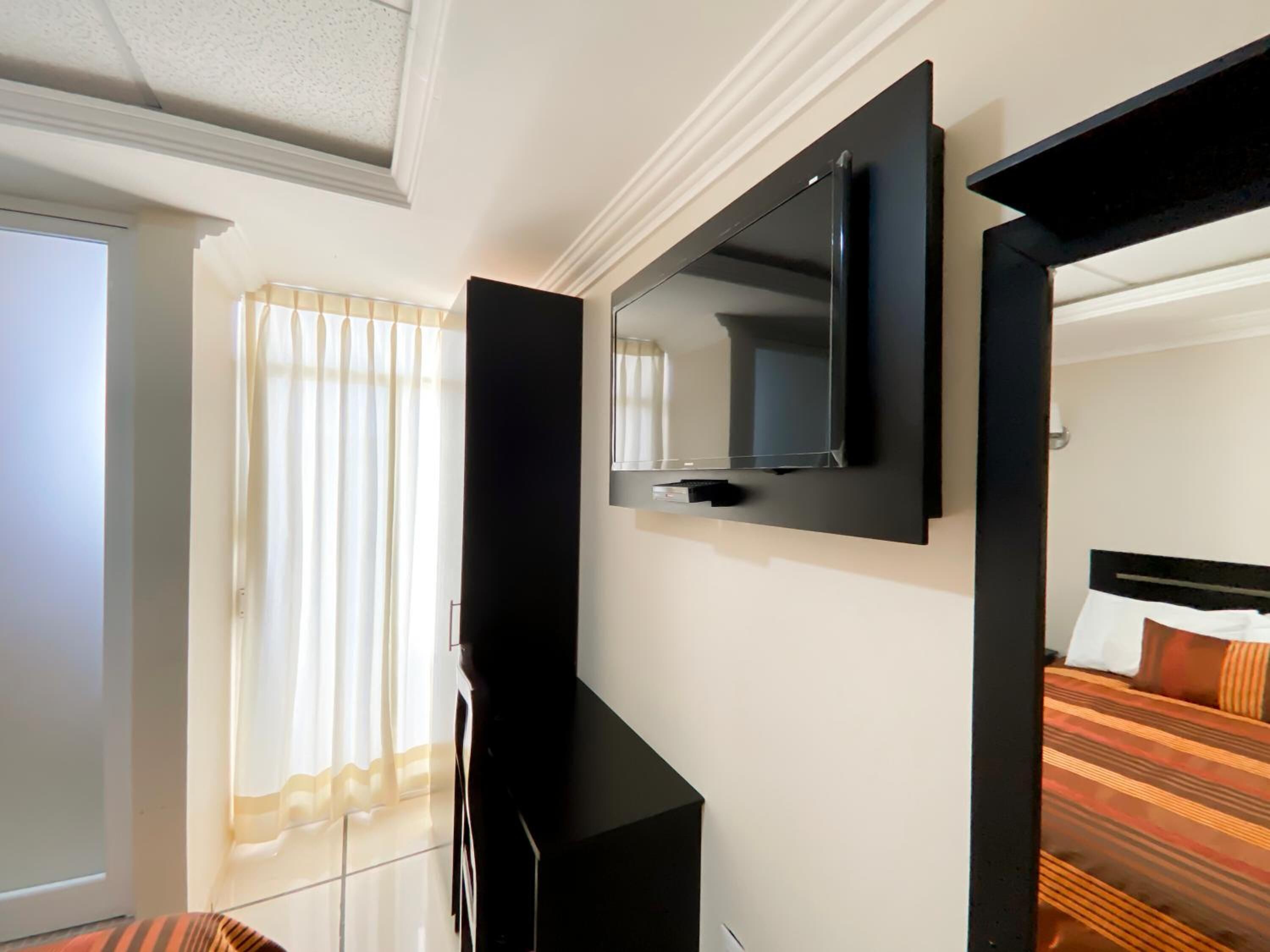A hotel room with a flat-screen TV mounted on the wall, a bed with striped bedding, and a window with sheer curtains.
