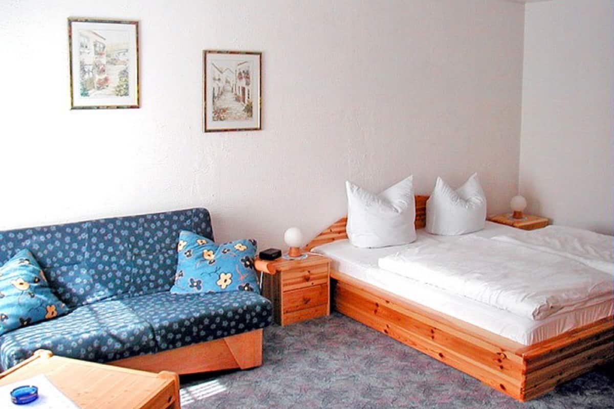Un dormitorio con una cama de madera, un sofá azul con cojines estampados, una pequeña mesa de madera y dos cuadros enmarcados en la pared.