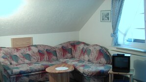 A corner living room with a floral sofa, a wooden coffee table, a television on a stand, and a window with curtains. - Gasthof Zum Dorfkrug (Winterberg)