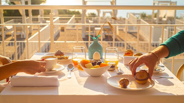 Una mesa dispuesta para el desayuno con vista al mar, con platos de comida, un tazón de fruta y vasos de jugo de naranja.