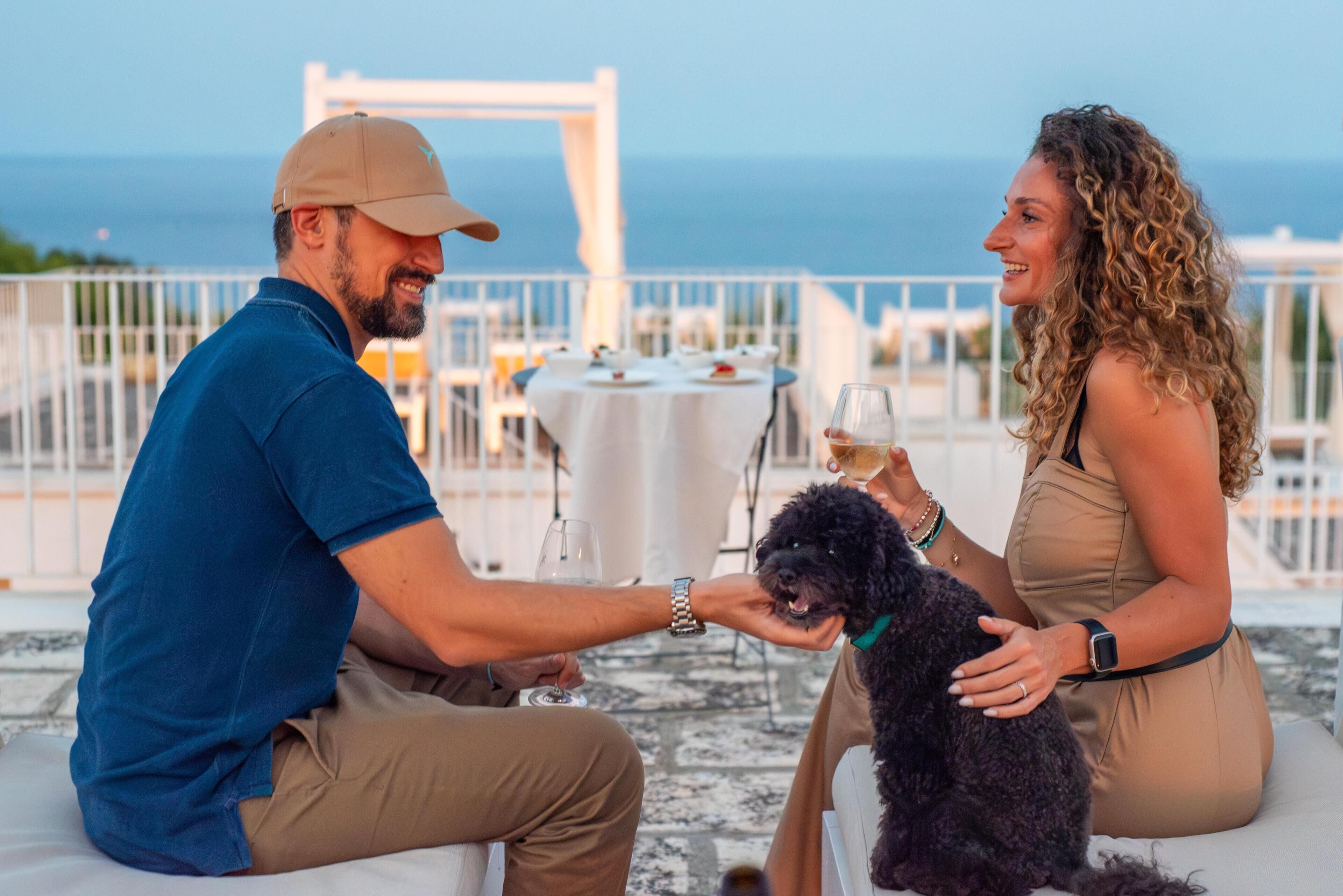 A couple seated at an outdoor table with a view of the sea, a dog, and a glass of wine.