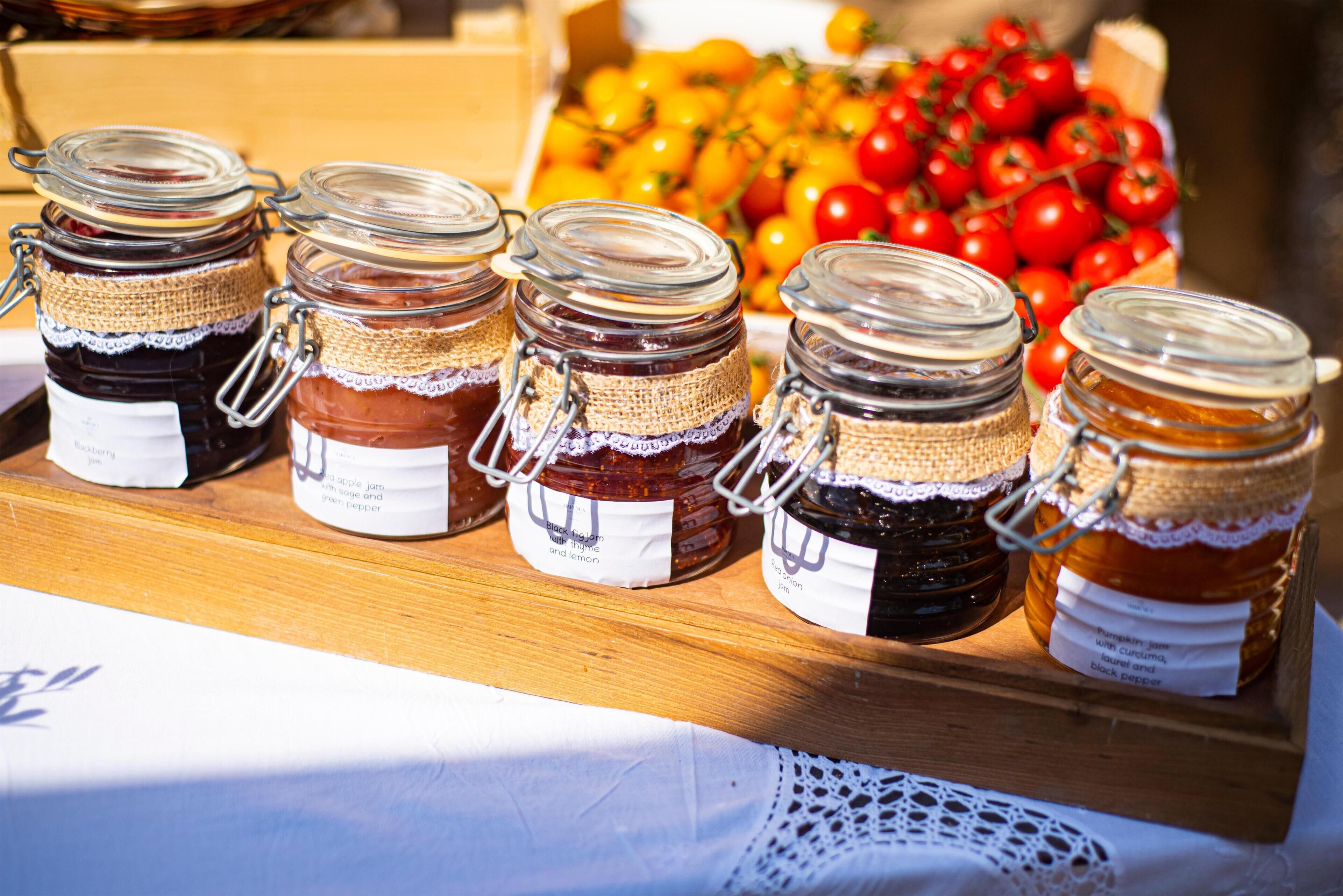 Eine Auswahl an Gläsern mit Etiketten, vermutlich mit verschiedenen Marmeladen oder Konfitüren gefüllt, auf einer Holzoberfläche mit weißem Tuch darunter.