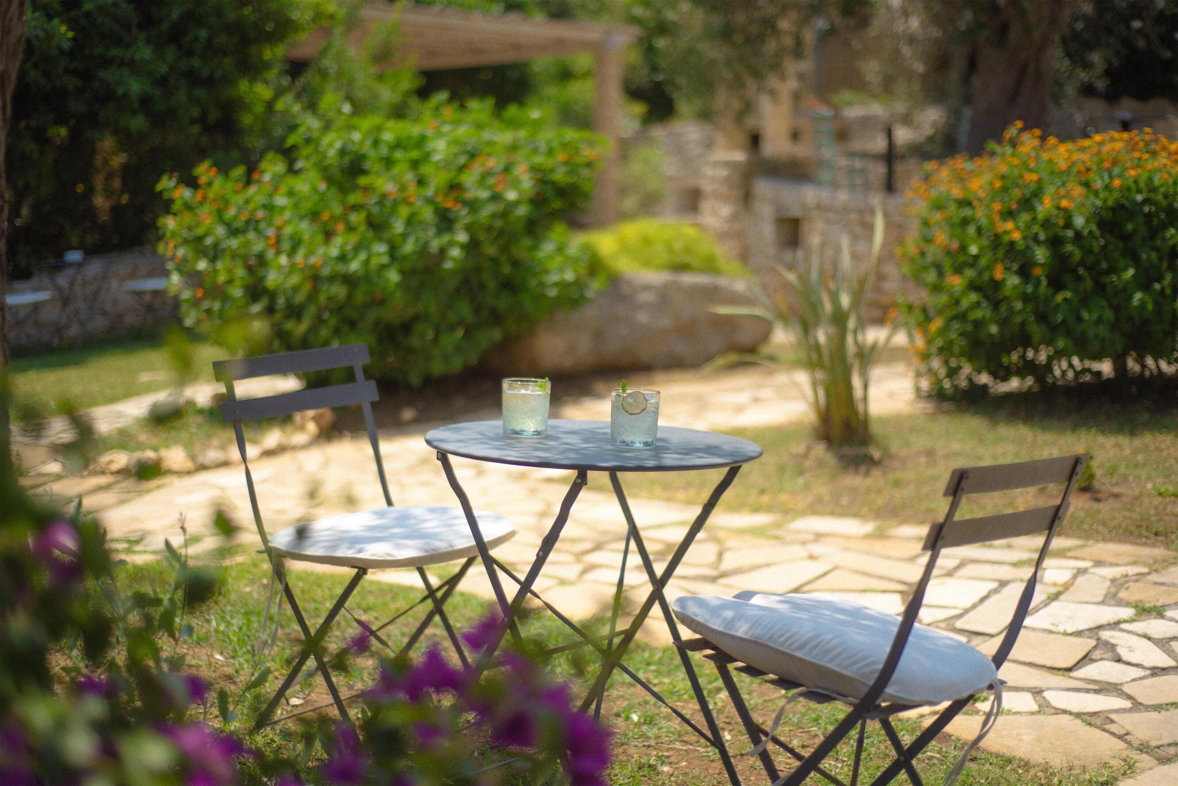 A garden patio with two chairs and a table, surrounded by greenery and stone pathways.