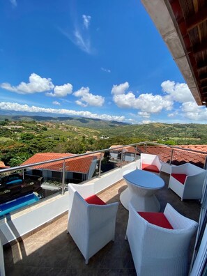 A balcony with white wicker furniture and a pool, overlooking a landscape with houses and hills. - Hotel Mansión Barichara (Barichara)