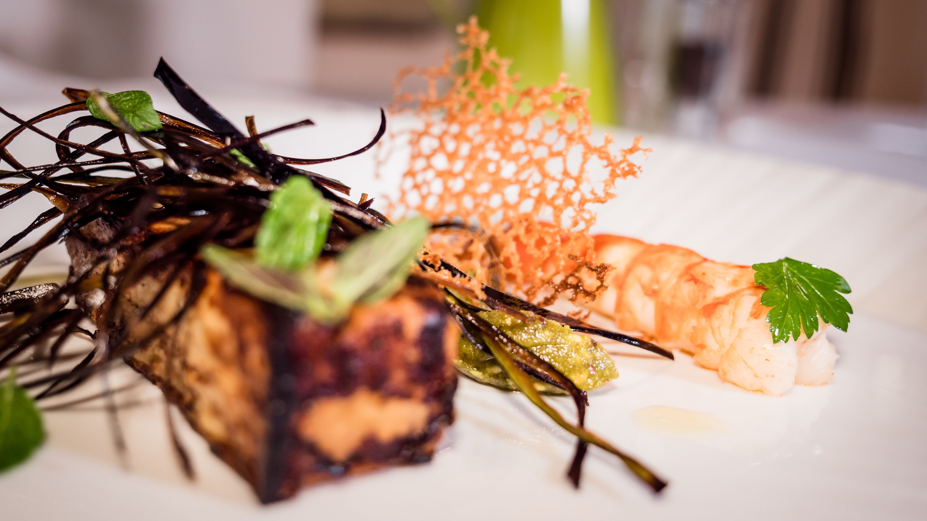 A plate of food with various dishes including a piece of meat, shrimp, and some garnishes.