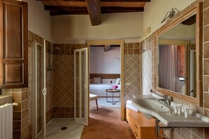 Un baño con ducha, lavamanos y una cama visible a través de una puerta abierta.