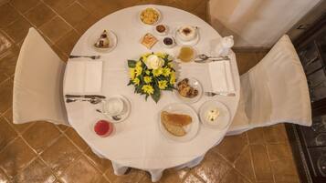 A round dining table with a white tablecloth, surrounded by chairs. There are plates with food, a vase of yellow flowers, and various condiments and utensils.