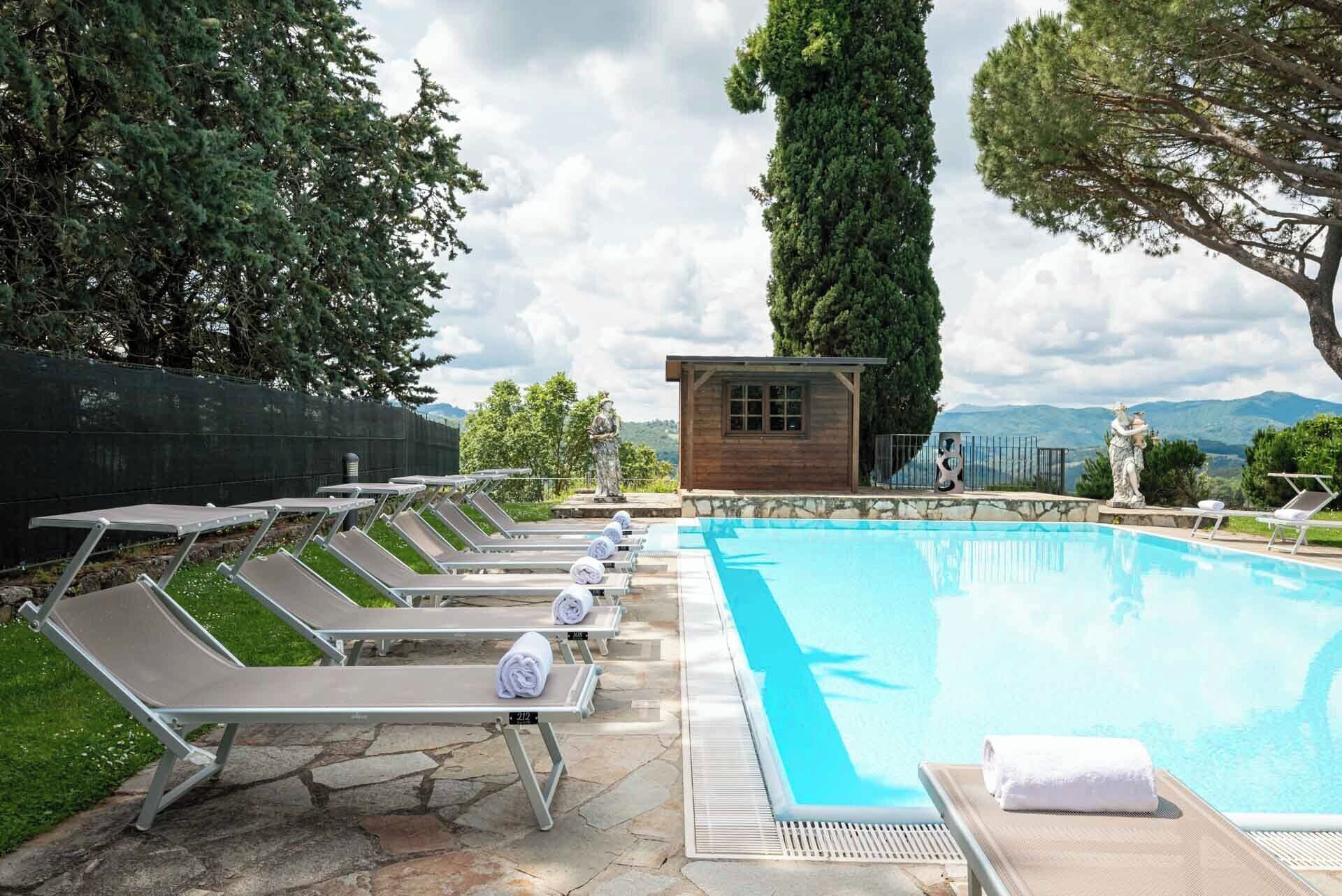 A swimming pool with lounge chairs and a small wooden building, surrounded by trees and a mountainous backdrop.