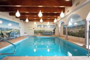 Una piscina interior con agua azul cristalina, vigas de madera en el techo y lámparas colgantes.