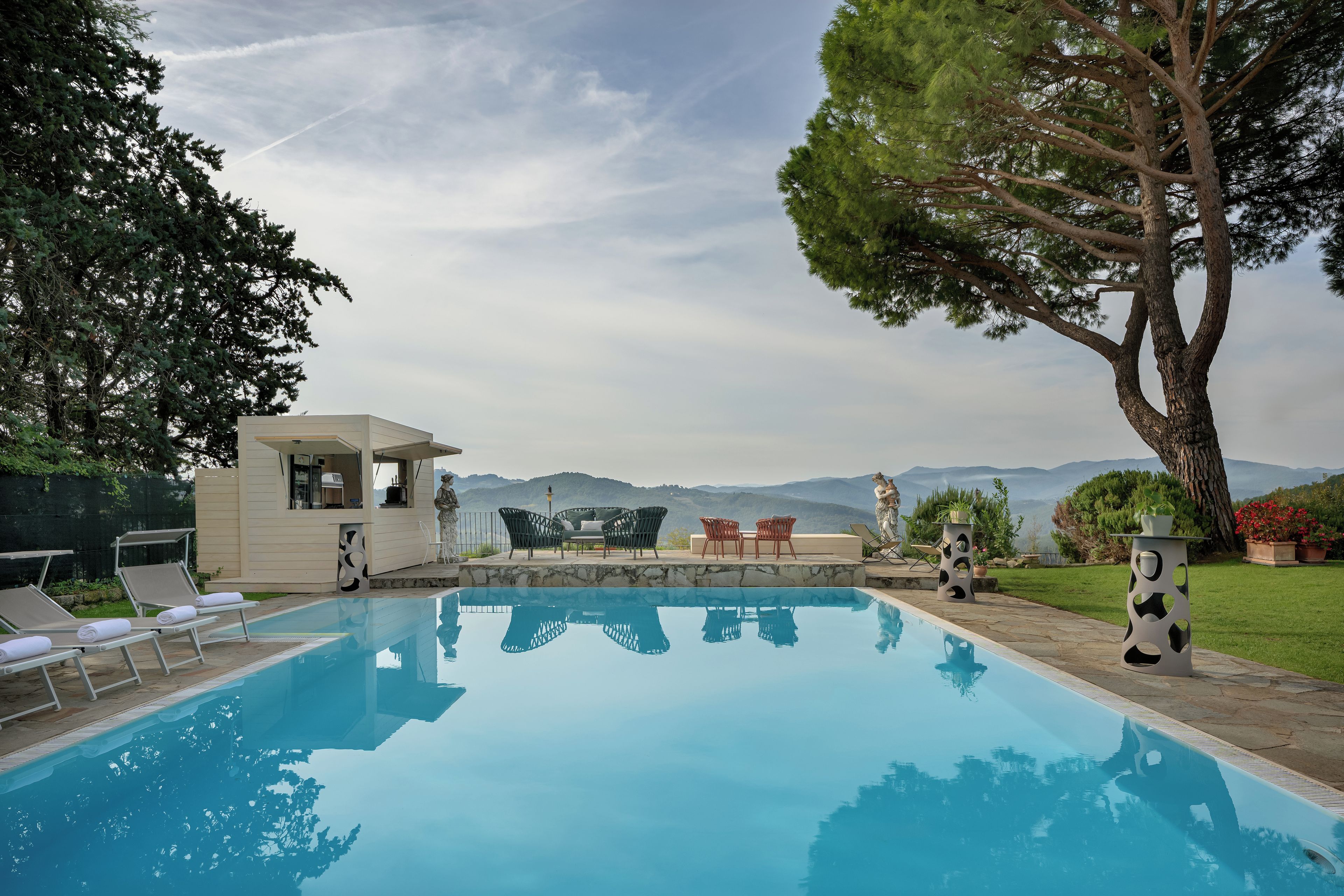 A swimming pool with lounge chairs and a table, surrounded by greenery and a view of distant mountains.