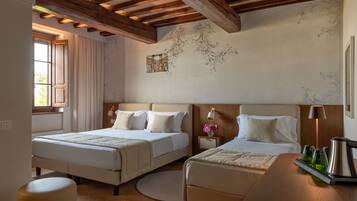A hotel room with two beds, wooden beams on the ceiling, a small table with a lamp, and a window with curtains.