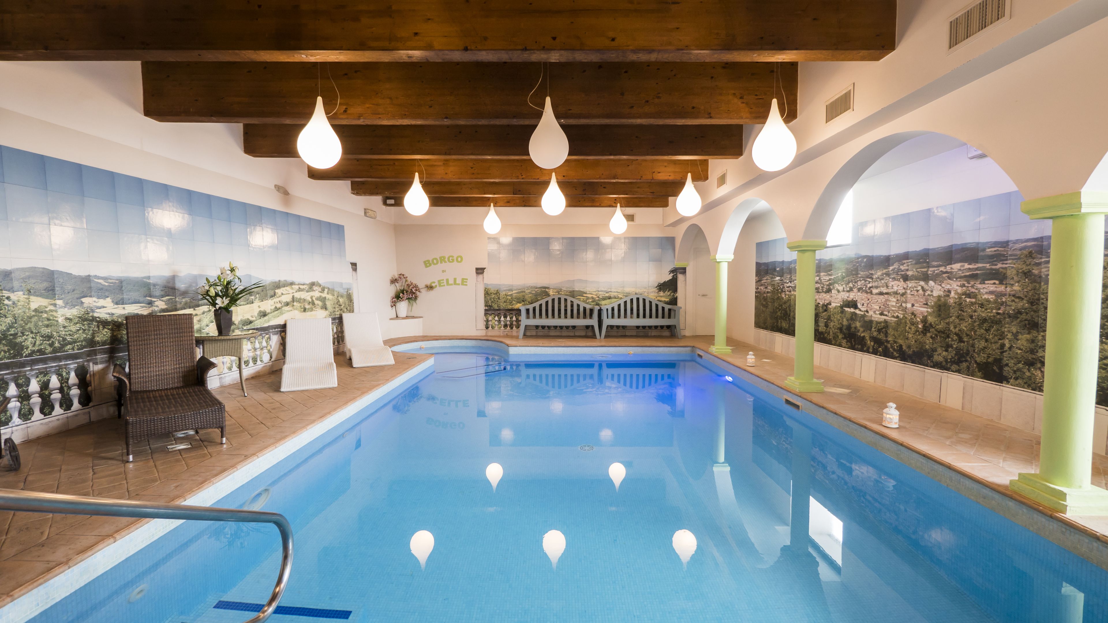 An indoor swimming pool with clear views of a mountainous landscape through large windows, wooden beams on the ceiling, and a few lounge chairs.