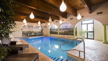 An indoor swimming pool with a view of a landscape through large windows, wooden ceiling beams, and a seating area with wicker chairs.