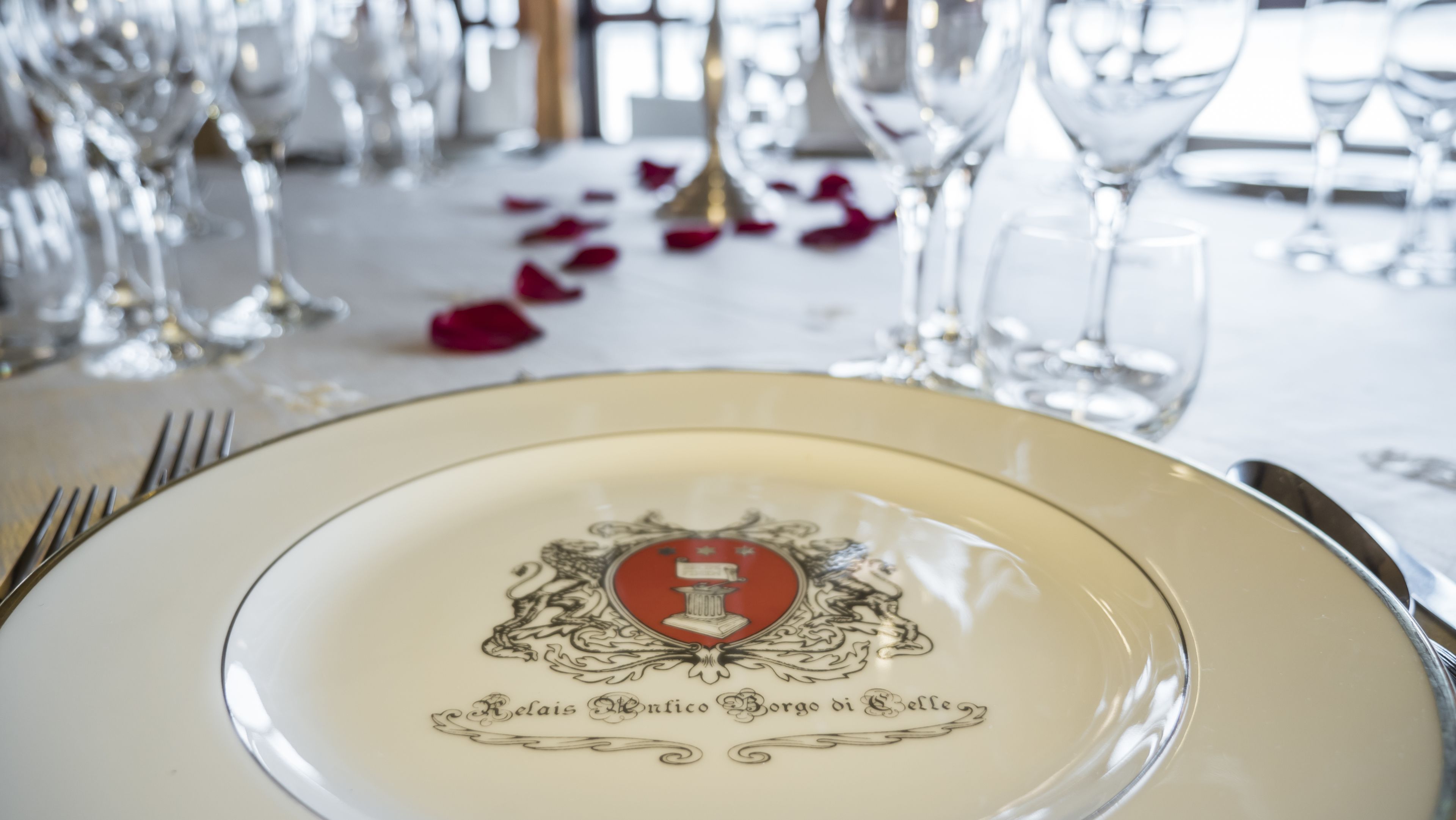 A table setting with a plate featuring an embossed emblem and text, surrounded by wine glasses and rose petals.