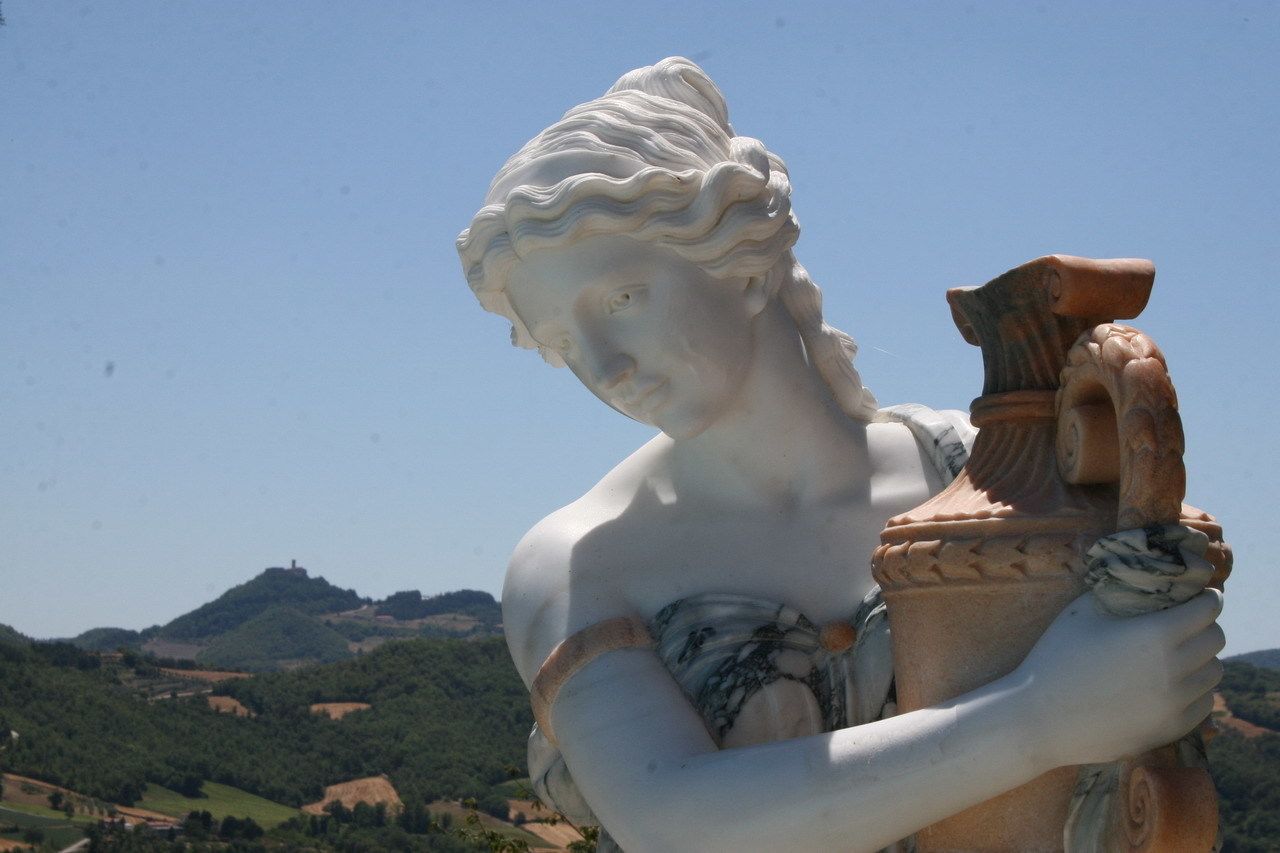 A statue of a classical figure holding a vase, set against a backdrop of rolling hills and a clear sky.