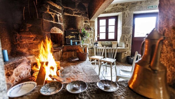 A rustic stone interior with a fireplace, wooden chairs, and a bell.