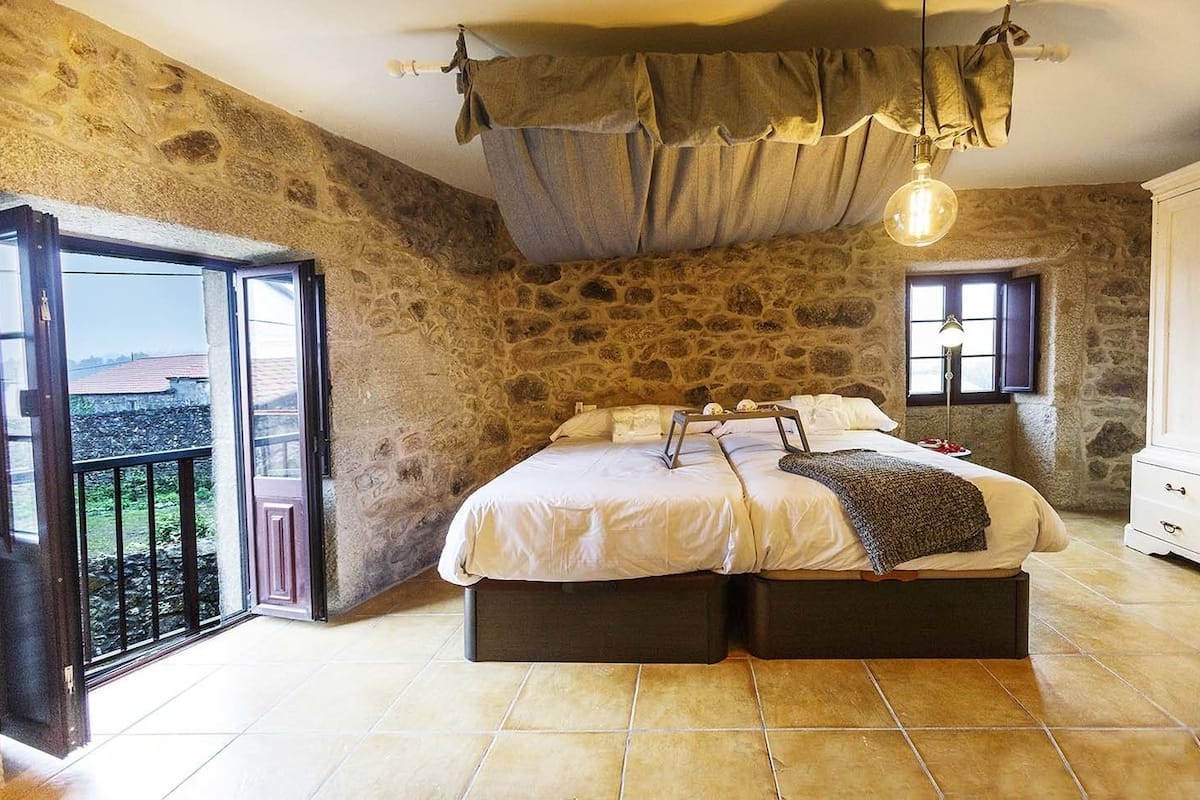 Un dormitorio con pared de piedra, una cama con cabecera, una ventana y un balcón.