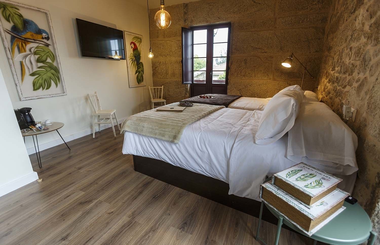 A bedroom with a bed, a small table with a coffee maker, a chair, and a TV on the wall.
