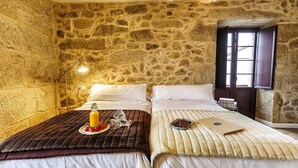 A room with two beds, stone walls, a window, and a bedside table with a bottle and glasses.