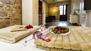 A cozy room with a stone wall, a bed with a fluffy blanket, a bedside table with a wine glass and a plate of fruit, and a wooden cabinet.