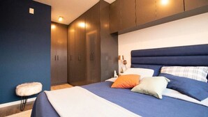 A modern bedroom with a large bed, dark wooden wardrobe, and a small white chair. - Apartamento em Santo Amaro (São Paulo)