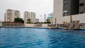 A swimming pool with lounge chairs and a view of high-rise buildings in the background. - Apartamento em Santo Amaro (São Paulo)