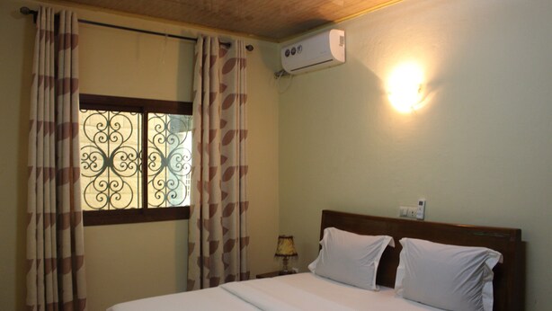 Une chambre d’hôtel comprenant un lit, des rideaux, une fenêtre ornée de ferronnerie décorative, un appareil de climatisation et une lampe de chevet.