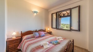A neatly made bed with a striped bedspread, two bedside lamps, a wooden nightstand, and a window with a view of greenery. - AffittaSardegna - Casa Pompia 1 (Siniscola)