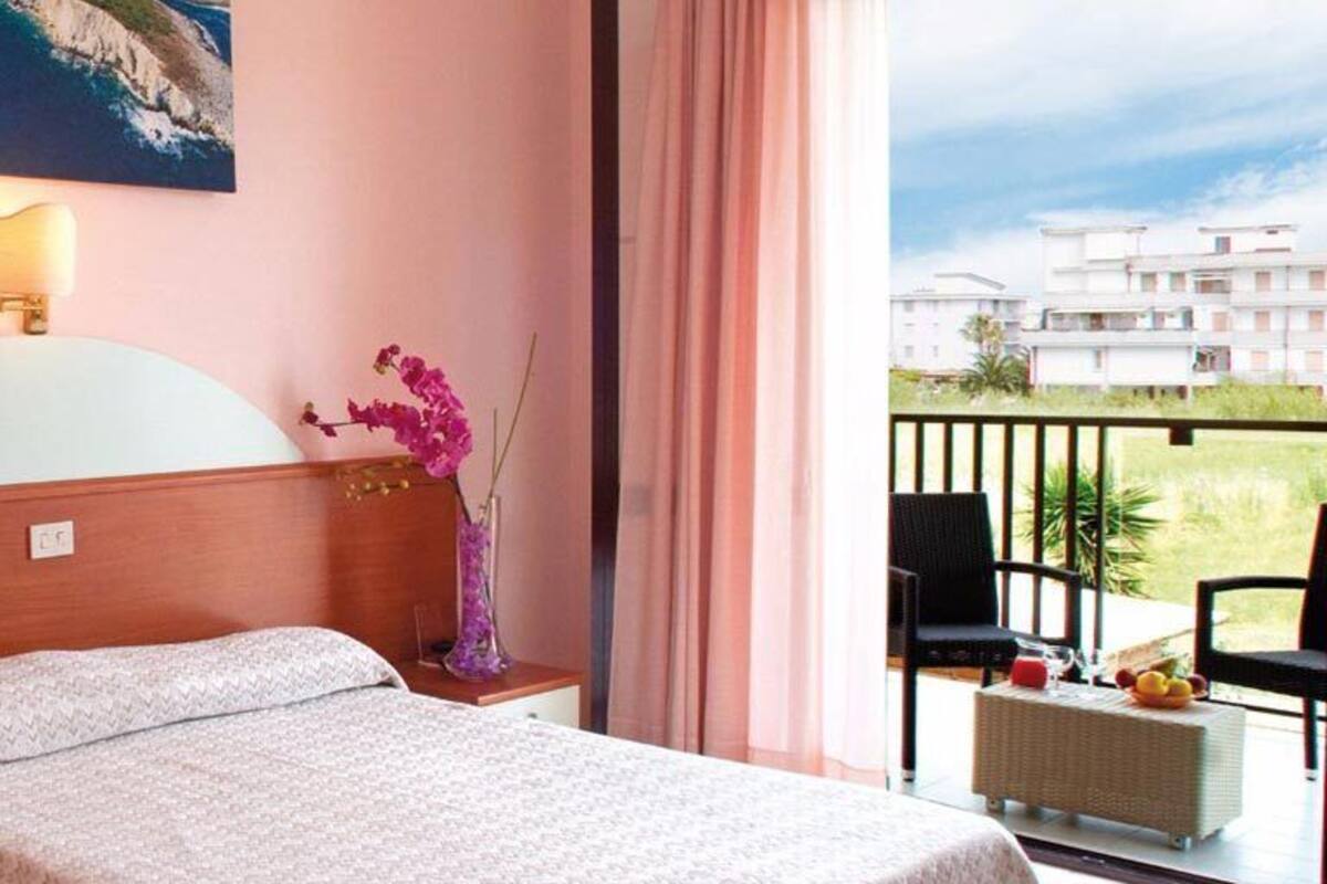 A hotel room with a bed, a nightstand, a vase with flowers, a balcony with chairs, and a view of buildings.