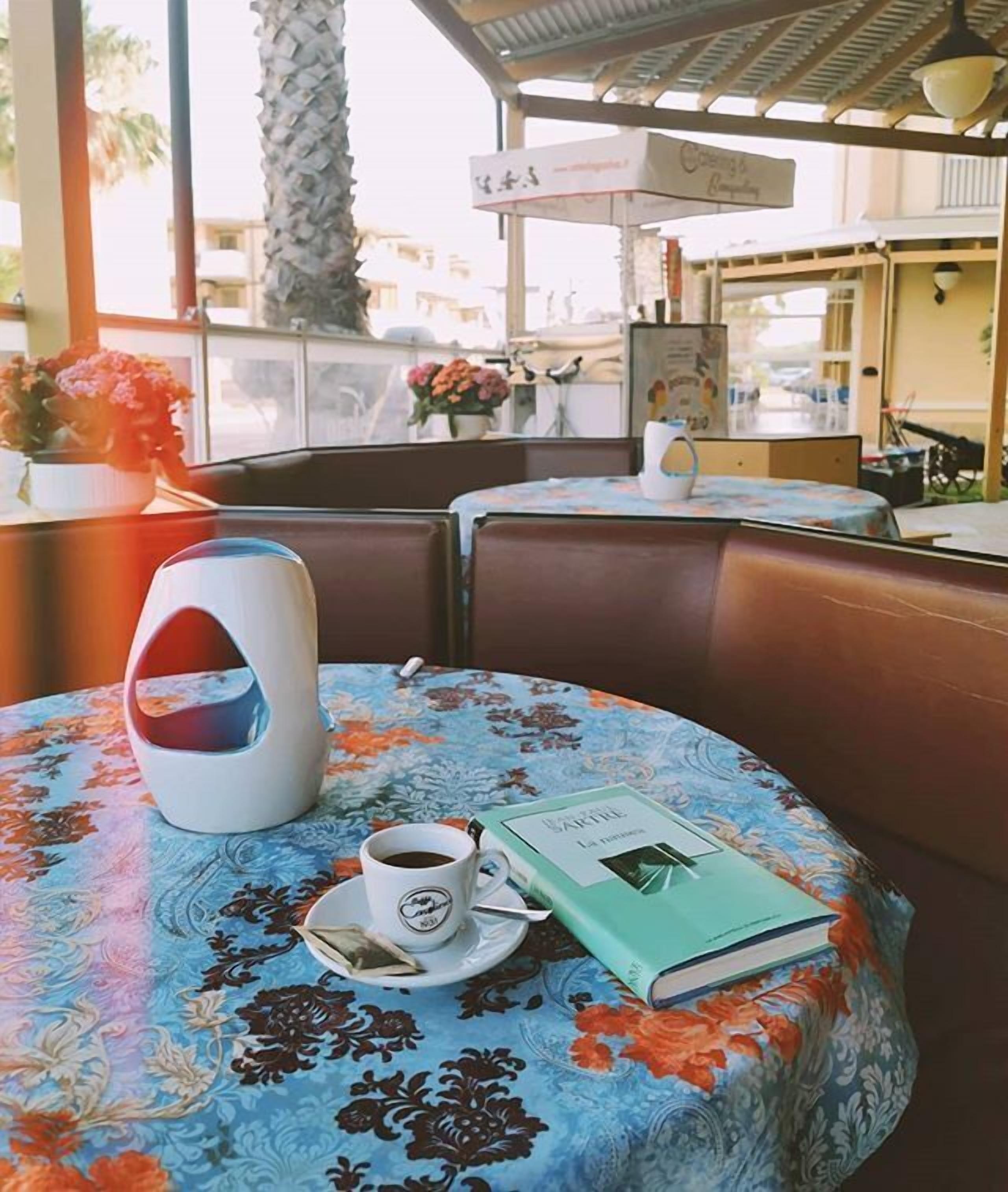 Un tavolo rotondo con tovaglia floreale, un teiera bianca, una tazza di caffè e un libro intitolato 'La romancita'.
