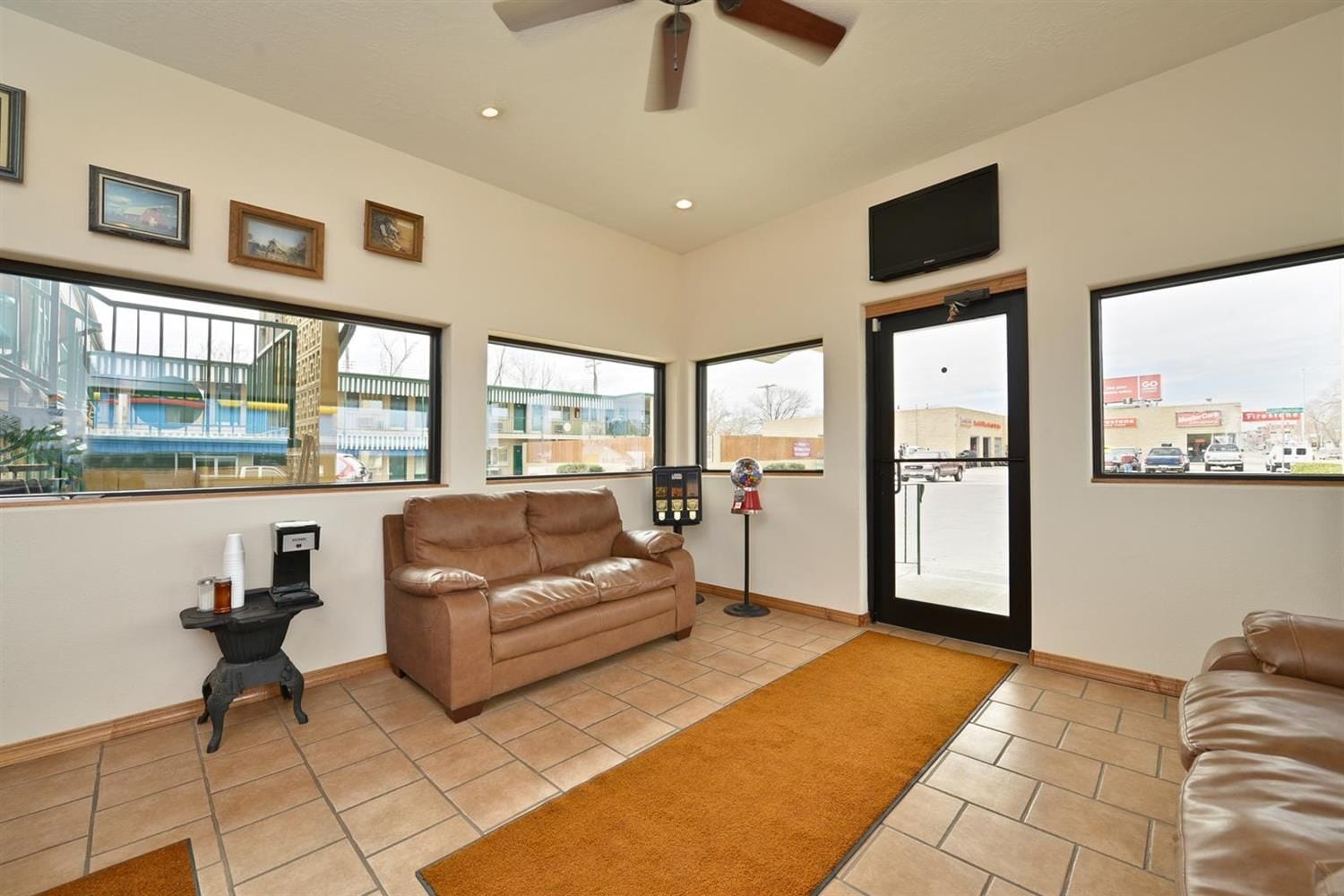 A spacious living room with a brown leather sofa, a coffee table, and a TV mounted on the wall. Large windows provide views of the outside.